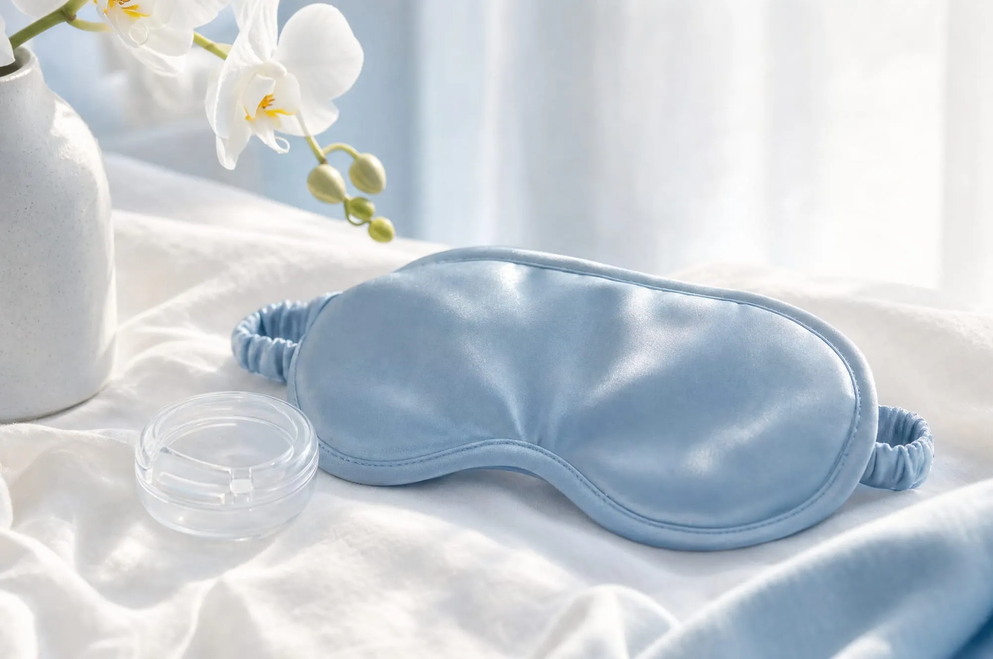 A silk sleep mask and clear retainer case on white bedside linen