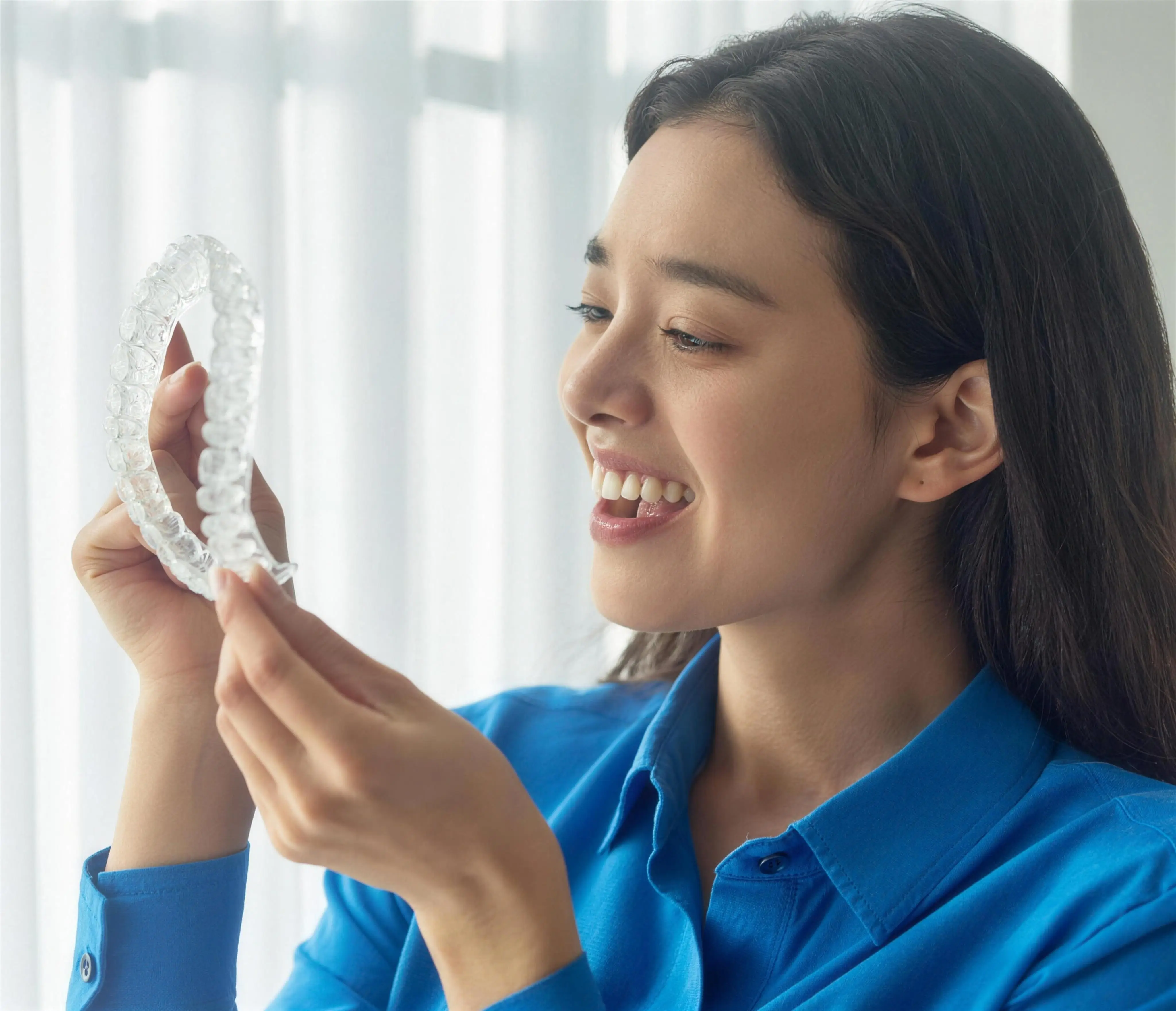 A smiling patient examining her clear aligner tray by a bright window