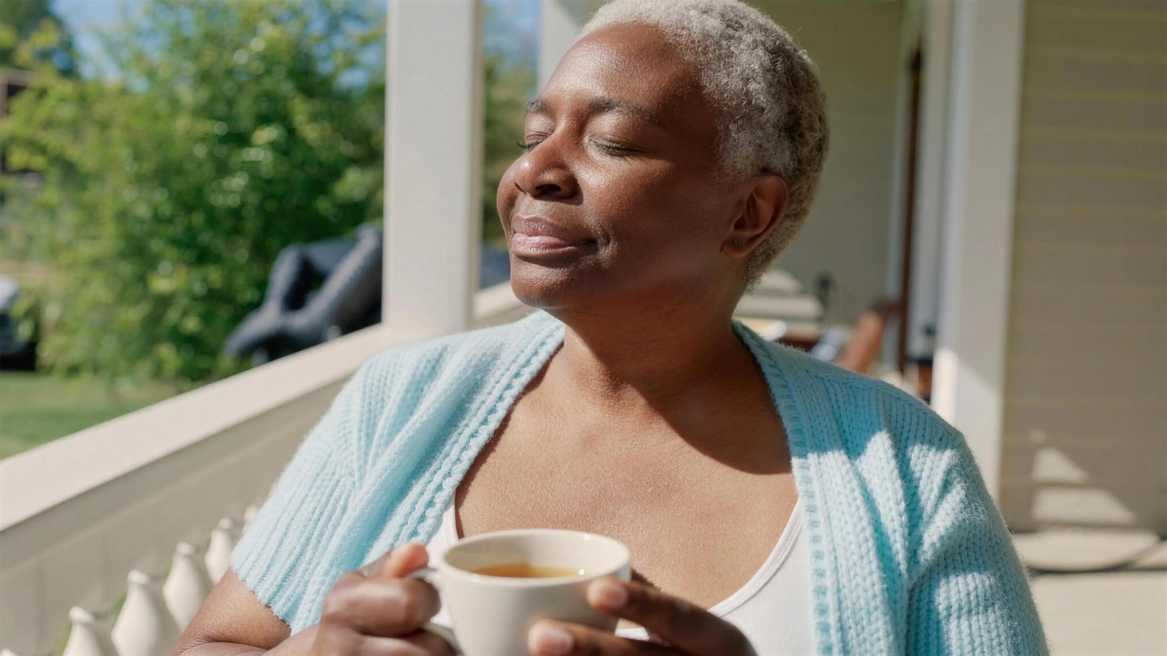 A woman smiling with closed eyes in the morning sun on her porch, tea in hand