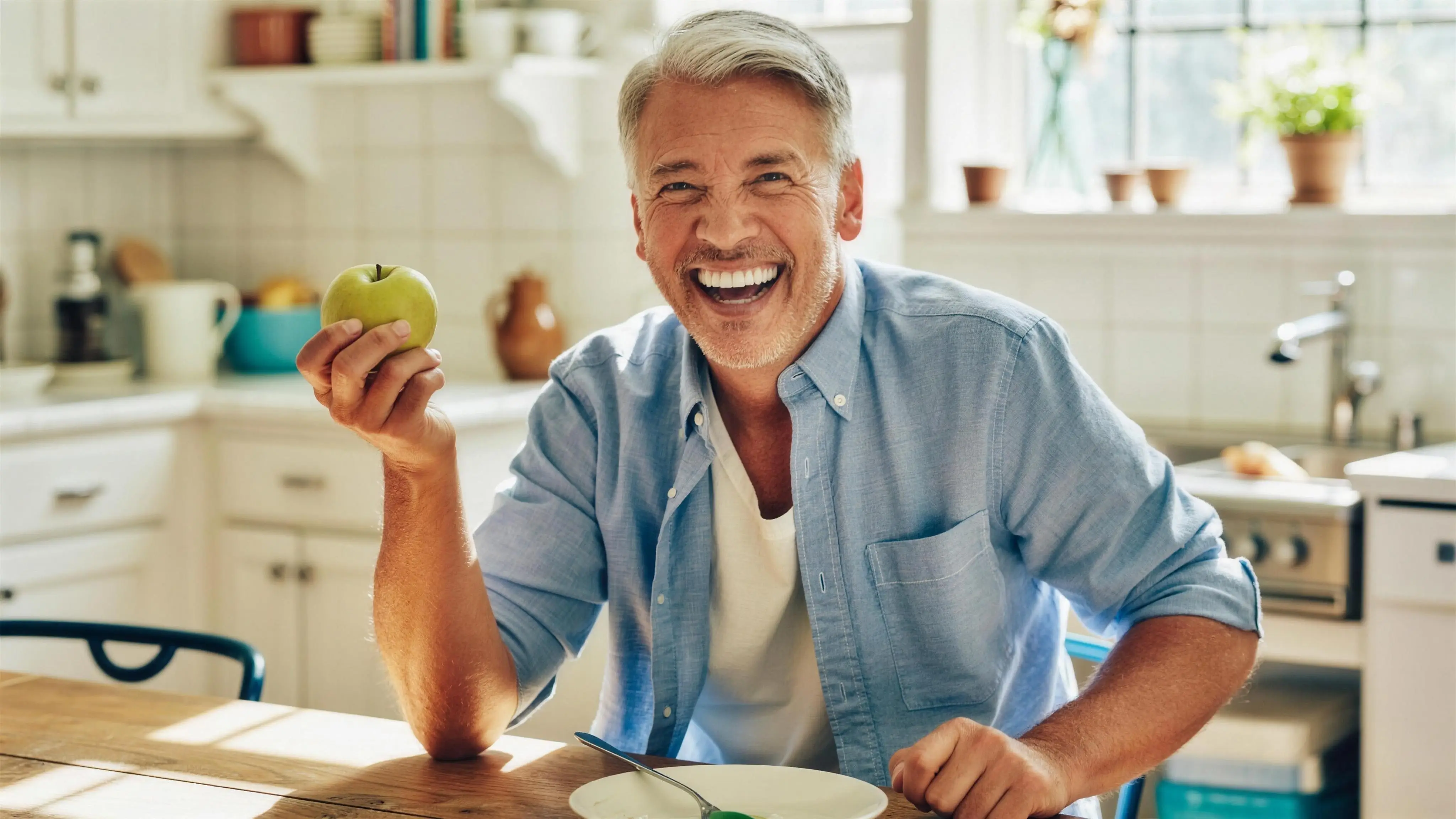 A man laughing at his sunlit kitchen table, apple in hand